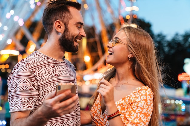 Afbeelding van een positief liefdevol stel dat buiten in het pretpark loopt met een mobiele telefoon.