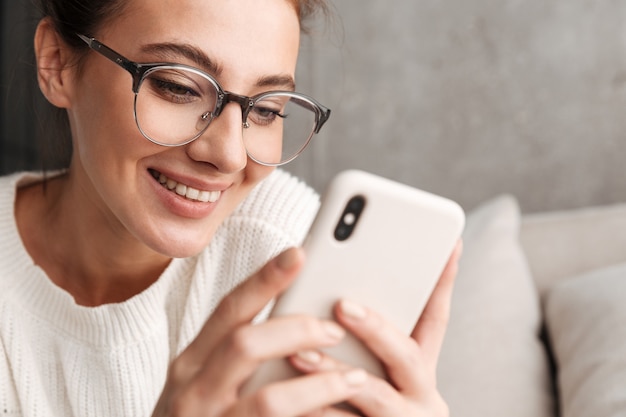 Foto afbeelding van een positieve brunette jonge vrouw in een bril die lacht en thuis een mobiele telefoon vasthoudt