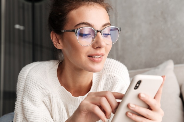 Afbeelding van een positieve brunette jonge vrouw in een bril die lacht en thuis een mobiele telefoon vasthoudt