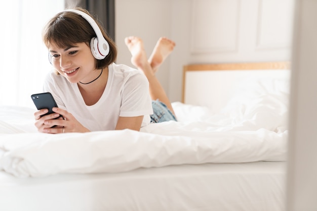 Afbeelding van een positieve gelukkige jonge mooie vrouw ligt binnenshuis thuis op bed en luistert naar muziek met een koptelefoon met behulp van een mobiele telefoon.