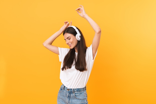 Afbeelding van een prachtige brunette vrouw met lang haar die lacht terwijl ze naar muziek luistert met een koptelefoon geïsoleerd over een gele muur