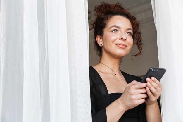 Afbeelding van een roodharige positieve optimistische jonge dame in lingerie badmode poseren binnenshuis in huishotel in de buurt van gordijn met behulp van mobiele telefoon.