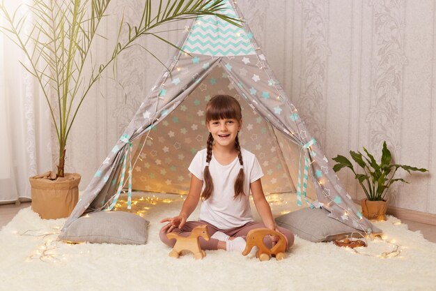 Afbeelding van een schattig gelukkig klein meisje met een wit t-shirt dat speelt met houten eco-speelgoed in een tipi charmant vrouwelijk kind met donker haar en vlechten die genieten van het spelen in haar nieuwe wigwam