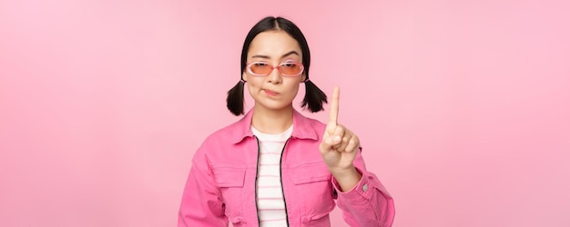 Afbeelding van een serieus, stijlvol aziatisch meisje met een zonnebril die een stop-verboden-gebaar-taboe-bord laat zien dat nee zegt over een roze achtergrond