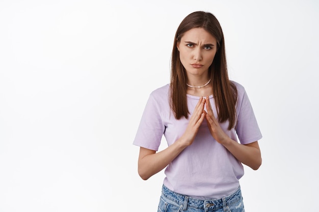 Afbeelding van een terughoudend grijnzend meisje dat een beledigd, mokkend gezicht maakt, vingers bij elkaar houdt en fronst, zich vastklampt en om iets vraagt, zich oneerlijk voelt, over een witte achtergrond staat