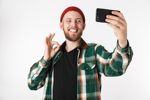 Afbeelding van een tevreden man met hoed en geruite overhemd selfie foto nemen op mobiele telefoon, terwijl geïsoleerd op witte achtergrond