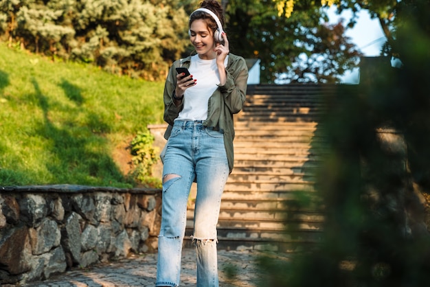 Afbeelding van een tevreden vrouw in een koptelefoon die naar muziek luistert met een smartphone tijdens het wandelen in het groene park