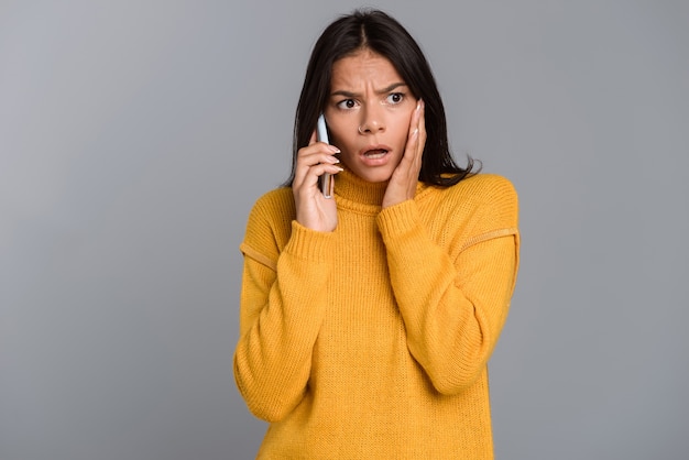 Afbeelding van een triest bang vrouw poseren geïsoleerd over grijze muur praten via de mobiele telefoon.