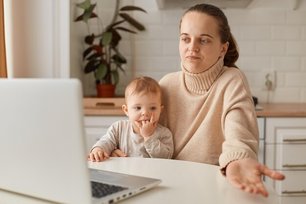 Afbeelding van een verbaasde donkerharige vrouw met een paardenstaart die een beige trui draagt die in de keuken poseert en een online gesprek heeft met een peuterdochter in handen die haar schouders ophaalt omdat ze niet zeker is