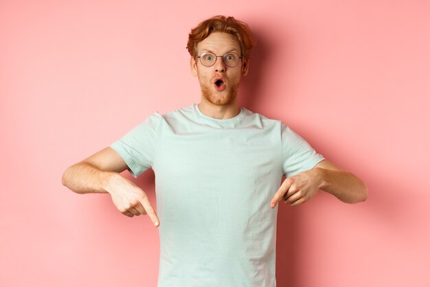 Afbeelding van een verbaasde jonge man met rood haar en baard met een bril en een t-shirt die met zijn vingers naar beneden wijst...