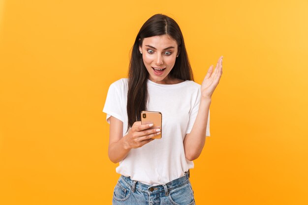 Afbeelding van een verraste brunette vrouw die eenvoudige kleding draagt die zich verheugt en een mobiele telefoon vasthoudt die over een gele muur is geïsoleerd