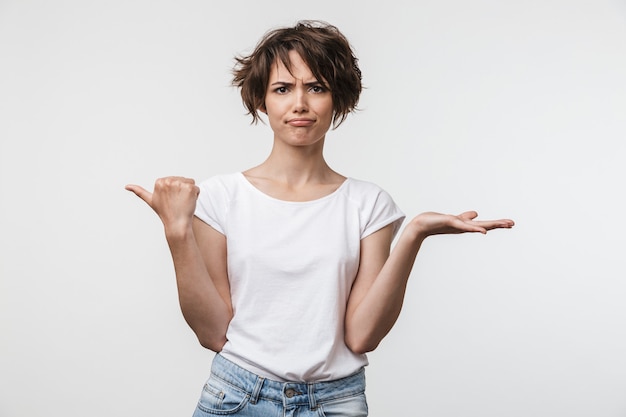 Afbeelding van een verwarde vrouw met kort haar in een basic t-shirt die met de vinger opzij wijst naar copyspace geïsoleerd over een witte muur