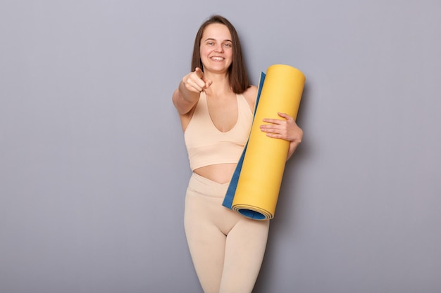 Foto afbeelding van een vriendelijke sportieve vrouw in een beige trainingspak met een yogamat geïsoleerd op een grijze achtergrond die naar je wijst terwijl je kiest voor gratis training in een nieuwe sportschool