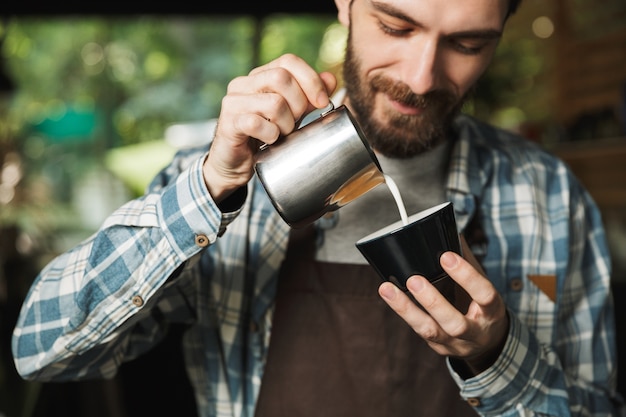 Afbeelding van een vrolijke barista-man met een schort die koffie maakt terwijl hij in een café of koffiehuis buiten werkt