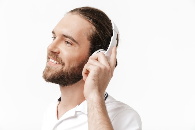 Afbeelding van een vrolijke gelukkige jonge knappe bebaarde man die zich voordeed over een witte muur die muziek luistert met een koptelefoon.