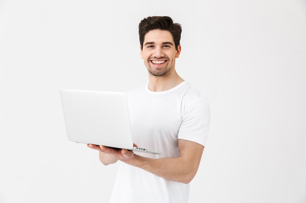 Afbeelding van een vrolijke opgewonden jongeman die zich voordeed op een witte muur met een laptopcomputer.