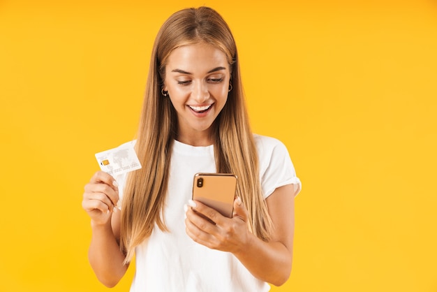 Afbeelding van een vrolijke vrouw in eenvoudige kleding die lacht terwijl ze een smartphone en een creditcard vasthoudt die over een gele muur is geïsoleerd