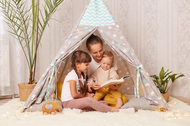 Afbeelding van een vrouw met haar kleine dochters die een boek lezen in een krot thuis Blanke vrouw zittend op de vloer in wigwam leest een sprookje voor aan haar kinderen die gelukkige tijd samen doorbrengen