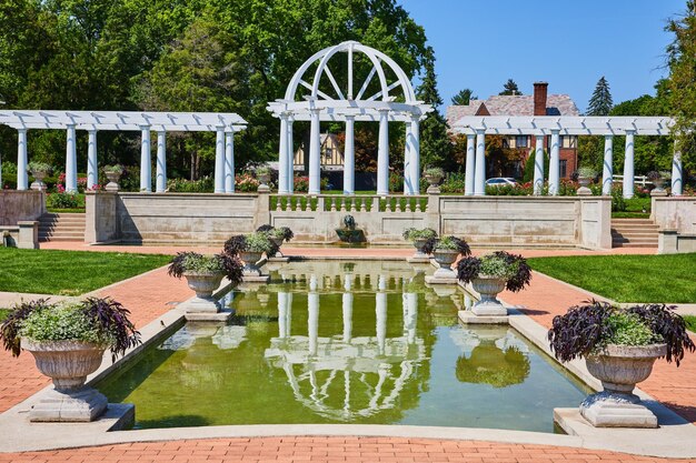 Afbeelding van een waterplas met stenen bloempotten die leiden naar de leeuwenkopfontein en witte pergola en hekjes