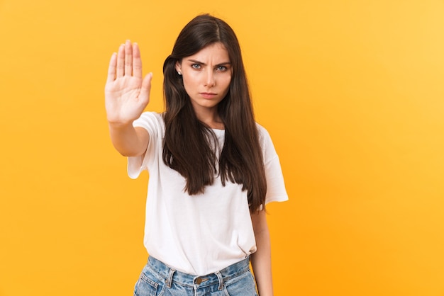 Afbeelding van een woedende vrouw van 20 met donker haar die fronst en een stopgebaar doet met palm geïsoleerd over gele muur