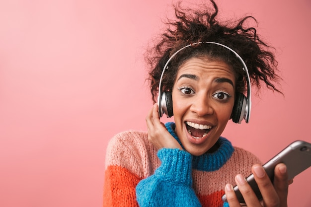 Afbeelding van emotionele mooie jonge Afrikaanse vrouw poseren geïsoleerde luisteren muziek met koptelefoon met behulp van mobiele telefoon.