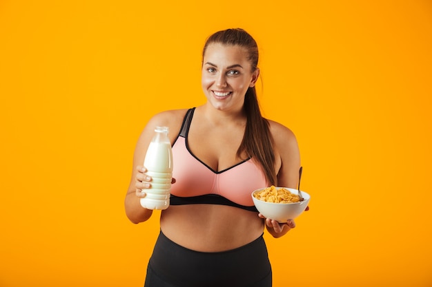 Afbeelding van europese mollige vrouw in trainingspak met melk en cornflakes voor ontbijt, geïsoleerd op gele achtergrond