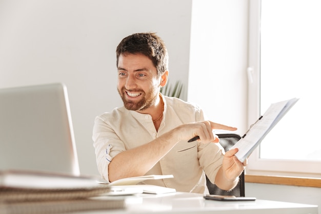 Afbeelding van gelukkig kantoormedewerker 30s dragen wit overhemd met behulp van laptop en papieren documenten, zittend aan tafel op de moderne werkplek