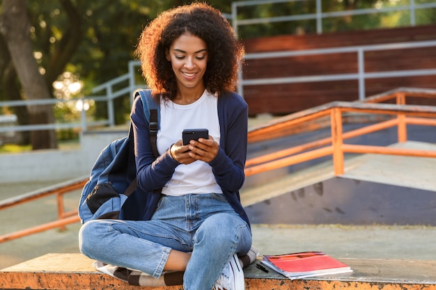 Afbeelding van gelukkige Afrikaanse jonge vrouw buiten zitten in park met behulp van mobiele telefoon.