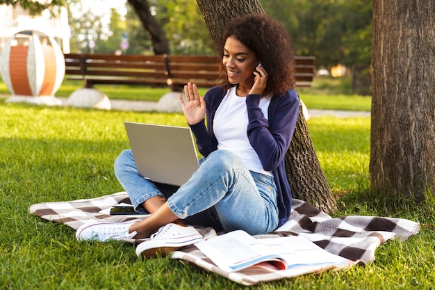 Afbeelding van gelukkige Afrikaanse jonge vrouw buiten zitten in park met laptopcomputer luisteren muziek met koptelefoon.