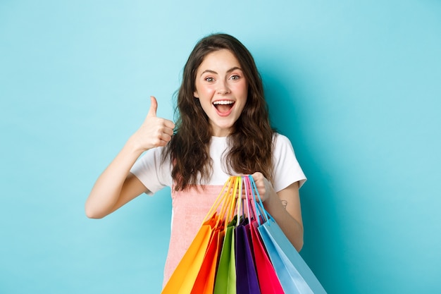 Afbeelding van gelukkige jonge glamour vrouw winkel in winkels met kortingen, duim opdagen, boodschappentassen vasthouden, opgewonden kijken naar camera, blauwe achtergrond.