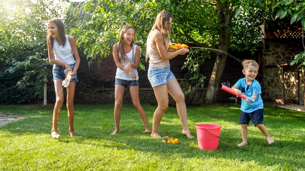 Afbeelding van gelukkige vrolijke kinderen met jonge moeder die speelt met waterpistolen en tuinhuis. Familie spelen en plezier hebben buiten in de zomer