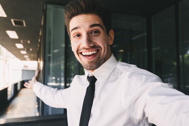 Foto afbeelding van gelukkige zakenman gekleed in formeel pak staande buiten glazen gebouw, en selfie foto te nemen