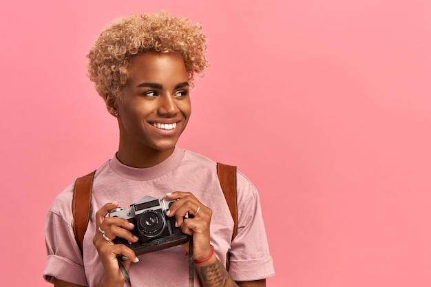Afbeelding van getalenteerde Afro-vrouwelijke fotograaf maakt professionele foto's tijdens avontuurlijke reis maakt gebruik van retro camera draagt lavendelt-shirt en rugzak geniet van vakantie poses binnenshuis Actief rustconcept