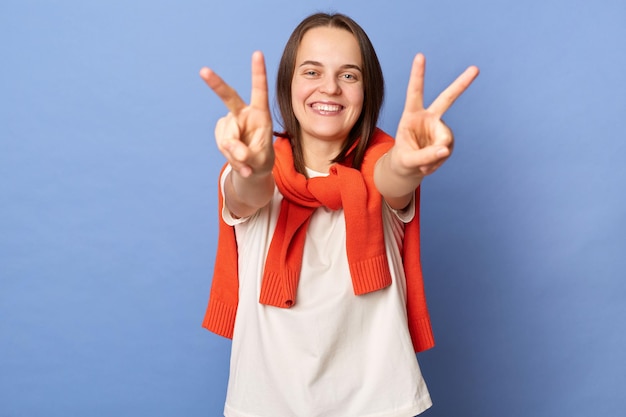 Afbeelding van glimlachende aantrekkelijke gelukkige blije vrouw gekleed in wit T-shirt en vastgebonden trui over schouders die overwinningsgebaar toont, toont v-teken staande geïsoleerd op blauwe achtergrond