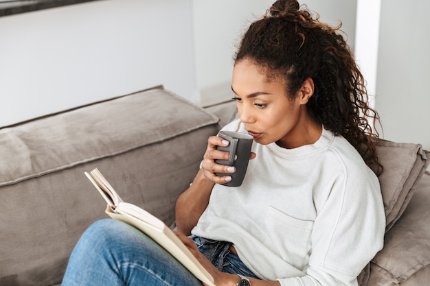 Afbeelding van jong Afrikaans Amerikaans meisje boek lezen en thee drinken, zittend op de Bank in lichte flat