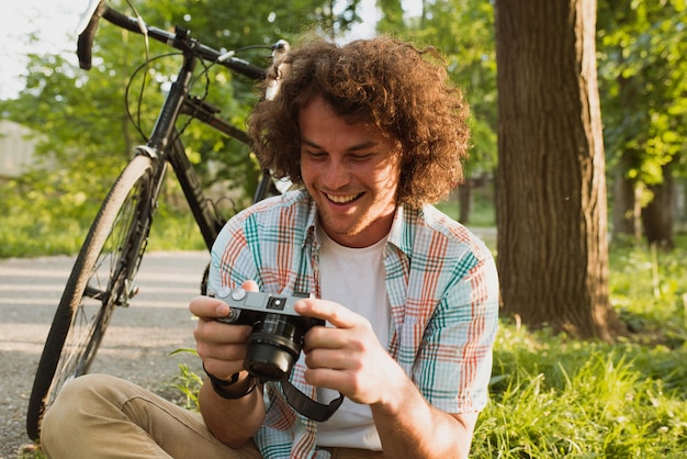 Afbeelding van jonge man die lacht met krullend haar op zoek naar foto op zijn digitale camera Knappe man draagt casual shirt ontspannen en zittend in de buurt van de fiets op de berg Mensen reizen en lifestyle concept