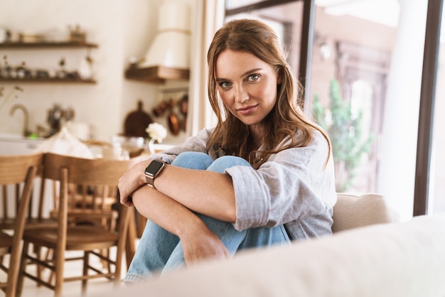 Afbeelding van jonge mooie roodharige vrouw binnenshuis zittend in de keuken thuis.