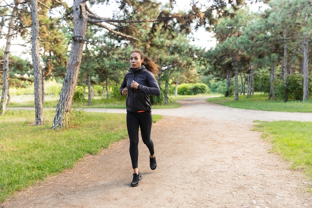 Afbeelding van jonge vrouw 20s dragen zwarte trainingspak uit te werken, terwijl het rennen door groen park