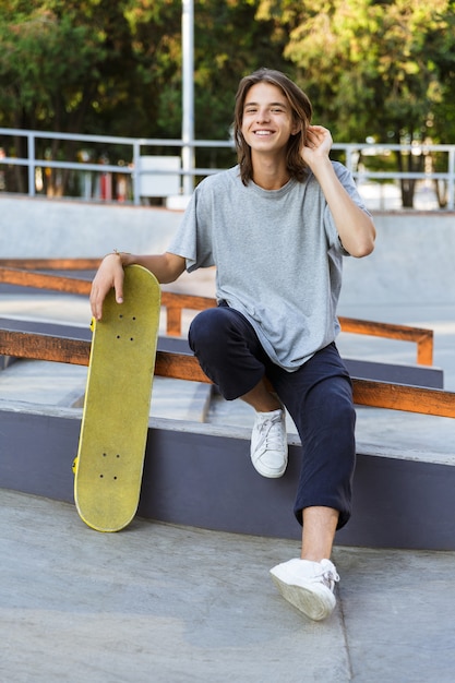 Afbeelding van knappe jonge skater man zitten in het park met skateboard.
