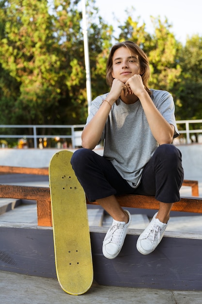 Afbeelding van knappe jonge skater man zitten in het park met skateboard.