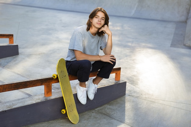 Afbeelding van knappe jonge skater man zitten in het park met skateboard.