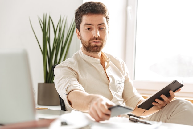 Afbeelding van knappe kantoormedewerker 30s dragen wit overhemd met behulp van smartphone en laptop, zittend aan tafel op de moderne werkplek