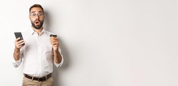 Afbeelding van knappe man die koffie drinkt en verrast reageert op een bericht op een mobiele telefoon die voorover staat