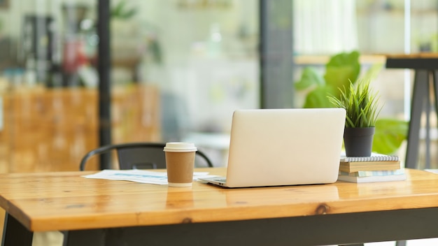 Afbeelding van laptopcomputerstandaard op houten tafel met koffiekopje, rapport, boeken, kamerplant in co-working space van de coffeeshop