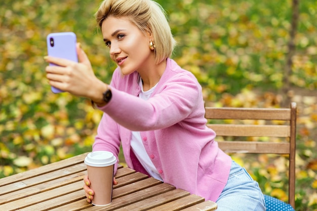 Afbeelding van mooie blonde jonge vrouw met blauwe spijkerbroek, witte t-shirt en roze jas glimlachend en selfie te nemen op smartphone Gelukkige vrouw die zelfportret maakt in het stadspark