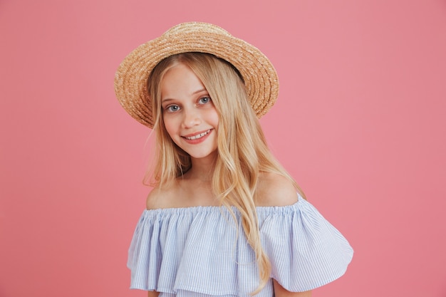 Afbeelding van mooie blonde zomer meisje 8-10 gekleed in een blauwe jurk en strooien hoed glimlachend in de camera met vrolijke blik, geïsoleerd op roze achtergrond