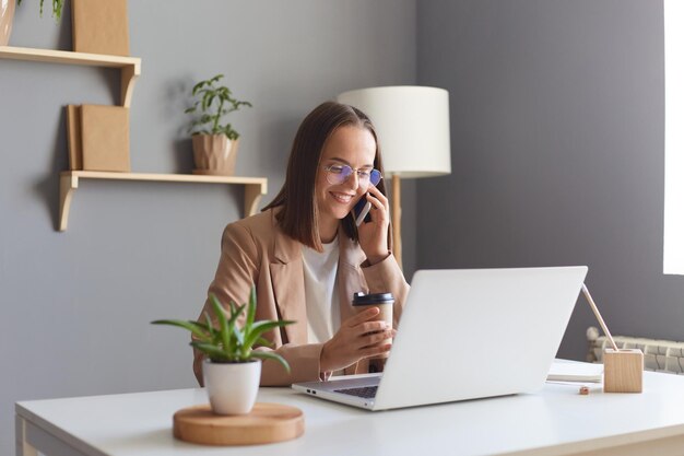 Afbeelding van mooie positieve aantrekkelijke jonge zakenvrouw die online werkt aan een notebook in het kantoor aan huis en praat op een mobiele telefoon die positieve emoties uitdrukt die koffie drinkt