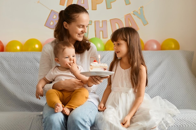 Afbeelding van opgewonden gelukkige kleine meisjes die op de bank zitten met moeder die zoete cake vasthoudt en glimlachend gelukkig verjaardag viert met mama en dochters zussen met moeder die in een goed humeur is