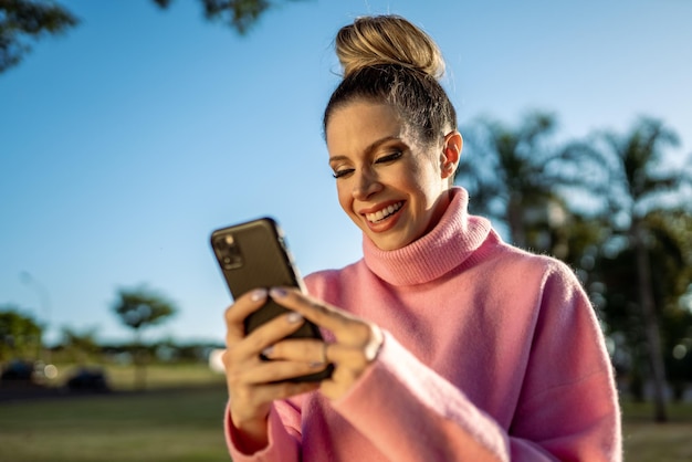 Afbeelding van opgewonden jonge blonde Latijnse vrouw die graag mobiel op parkachtergrond gebruikt