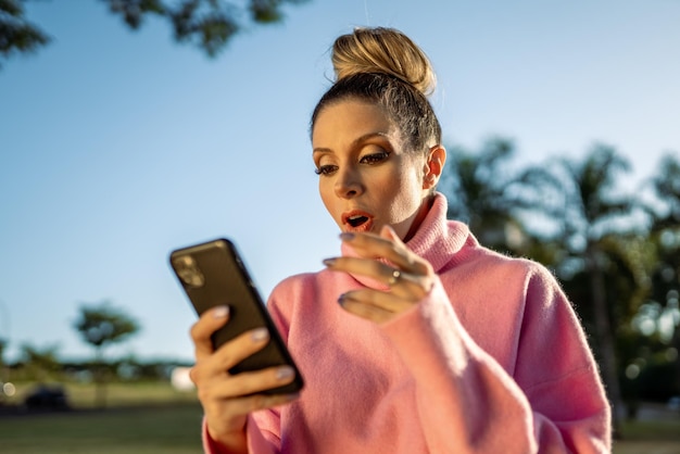 Afbeelding van opgewonden jonge blonde Latijnse vrouw verrast met behulp van mobiele telefoon op parkachtergrond